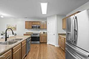 A modern kitchen with wood cabinets, stainless steel appliances, a double sink with a black faucet, a pantry with a white door, and hardwood flooring under bright overhead lighting.