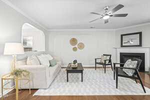 A living room with a beige sofa, two dark wood chairs, a black coffee table, a light area rug, a round side table with a lamp, a fireplace with a mirror above it, and three woven wall hangings on a light gray wall.