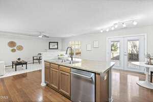 A modern open-concept kitchen with a central island and stainless steel dishwasher, overlooking a living area and a dining nook beside glass doors leading to a deck. The space features hardwood floors and neutral decor.