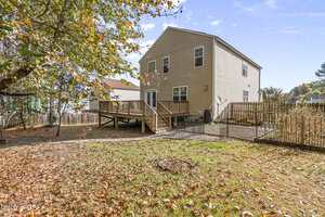 A tan two-story house with a wooden deck and fenced backyard, surrounded by trees. The yard has patchy grass and scattered leaves, with a gravel pathway leading from the deck.