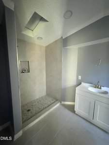 A bathroom with a large walk-in shower on the left, featuring light-colored tiles and a recessed shelf, and a white vanity with a sink and faucet on the right. The walls are painted gray.
