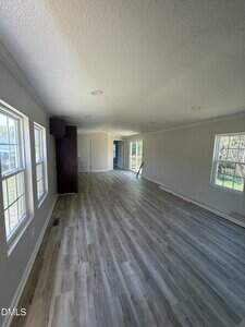 A long, empty room with light-colored wood flooring, beige walls, and several large windows along the left wall. There is a dark cabinet and a door at the far end, with natural light filling the space.