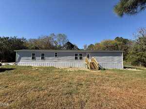 A single-story, light gray manufactured home at 485 Lemay Lane, Henderson features wooden steps leading to the front door. The home sits on a grassy lot with scattered trees and a clear blue sky overhead.