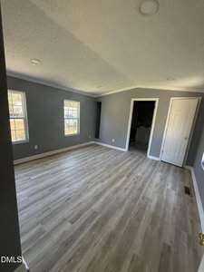 A room with gray walls, two rectangular windows, light wood flooring, recessed ceiling lights, and an open doorway leading to a bathroom. A white door is closed on the right side of the room.