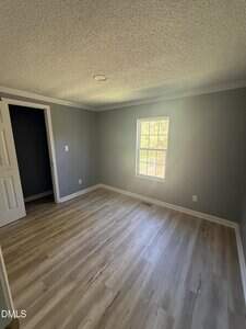 An empty room with light wood flooring, gray walls, a single window letting in natural light, a white door partially open, and white baseboards and ceiling trim.