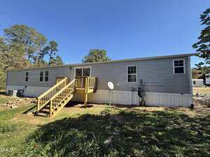 A single-story gray mobile home with white trim, multiple windows, a yellow wooden stairway and porch, a satellite dish, and utility meters, set on a grassy lot with trees in the background.