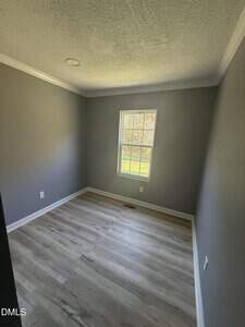 A small empty room with gray walls, white trim, light wood flooring, and a single window letting in natural light. The ceiling has a textured finish with a recessed light fixture.