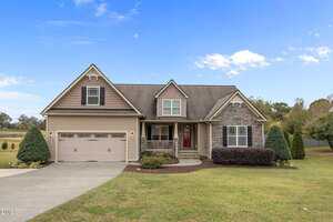 A suburban house in Wake Forest with a two-car garage, stone and siding exterior, covered front porch, and manicured lawn under a partly cloudy sky at 3522 Lavender Lane. Shrubs and trees are beautifully landscaped around the property.