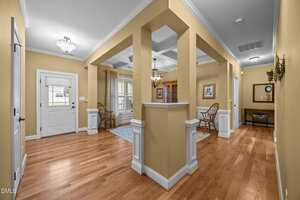 A hallway with light yellow walls, hardwood floors, white trim, and crown molding. The entry door is on the left, and wooden chairs, framed art, and a mirror decorate the space. Multiple doorways lead to other rooms.