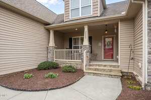 A front view of a house entrance with beige siding, stone accents, a covered porch with railing, and a small landscaped area with mulch and shrubs. The house number 3522 is displayed on a porch column.