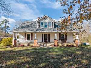 A white, two-story house at 595 George Sherman Road in Rougemont features a covered front porch with brick columns, a gabled roof, and a wooden porch swing. The grassy lot is dotted with trees, fallen leaves, and a propane tank in the background.