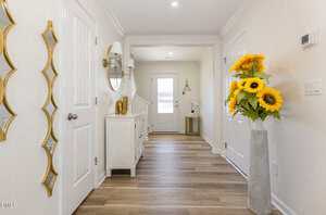 A bright hallway with light wood flooring, cream walls, a white front door, a sideboard with a round mirror and wall lamps, a decorative gold wall piece, and a tall vase of sunflowers.