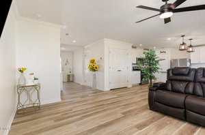 A modern living area with light wood flooring, white walls, a dark brown recliner, a gold accent table with decor, artificial plants, and a view into a white kitchen with pendant lights and a double-door pantry.