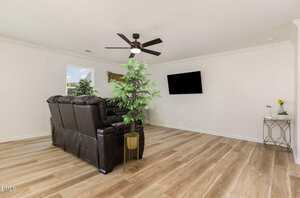 A living room with light wood flooring, a dark brown leather sofa, a ceiling fan, a wall-mounted TV, a potted plant, a small side table with decor, and a window letting in natural light.