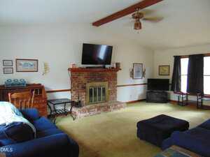 A living room with a brick fireplace, a TV mounted above it, a wooden desk to the left, a blue sofa and ottoman, two windows with dark curtains, and framed pictures on white walls.