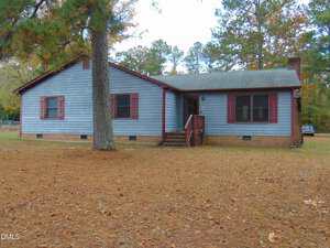 A single-story house with blue horizontal siding, maroon shutters, and a brick foundation sits on a pine needle-covered yard with tall trees in the background. Steps lead to a small front porch and door.