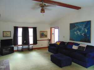 A living room with a dark blue sectional sofa and ottoman on a light green carpet. There are paintings on the walls, a ceiling fan, two windows with dark curtains, and a TV in the corner on a small stand.