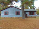 Charming single-story house at 111 E F Street, Butner, with gray wood siding, red shutters, and a brick foundation. A large pine tree and pine-needle-covered yard welcome you to this inviting home with steps leading to the front door.