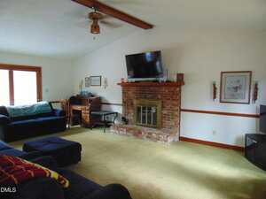 A living room with a brick fireplace, mounted TV above, and a brass fire screen. There is a blue sectional sofa, a wooden desk, framed art on the walls, and beige carpeted floor. A ceiling fan hangs from a beamed ceiling.