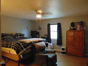 A bedroom with a bed against the left wall, black and white checkered bedding, a recliner chair in front, a wooden dresser to the right, and a window with dark curtains in the center of the back wall.
