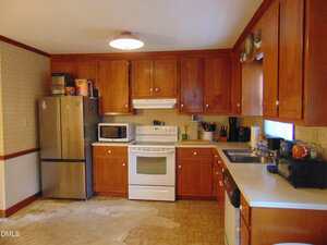 A kitchen with wooden cabinets, white countertops, a white stove and oven, a stainless steel refrigerator, a microwave, various small appliances, and a window above the sink. The floor shows visible discoloration.