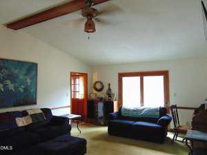 A living room with two dark blue couches, a wooden rocking chair, a ceiling fan with a light, a large painting of birds on the wall, and glass doors letting in natural light. A side table and small cabinet are also visible.