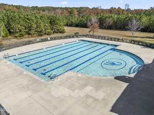 Large outdoor swimming pool with swim lanes and a circular logo on the pool floor, surrounded by concrete decking and black metal fencing, bordered by trees and open grassy areas. No people are present.