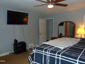 A bedroom with a checkered bedspread, a wooden dresser with a mirror, a TV mounted on the wall, a ceiling fan with lights, a floor lamp, and a small shelving unit beneath the TV.