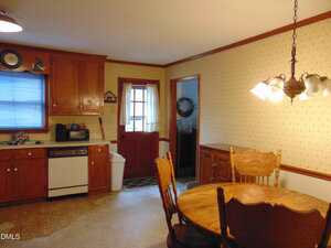 A kitchen with wooden cabinets, a dishwasher, a white trash can, and a round wooden dining table with chairs. The room has patterned wallpaper, a hanging light fixture, and a door with a window leading outside.