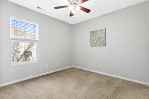A small, empty room with beige carpet, light gray walls, a window with white blinds, a ceiling fan with lights, and a single abstract painting hanging on one wall.