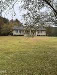 A single-story beige house with white trim and a red front door sits in the center of a large grassy yard at 3105 Tump Wilkins Road in Stem, surrounded by trees under an overcast sky. Branches partially frame the view in the foreground.