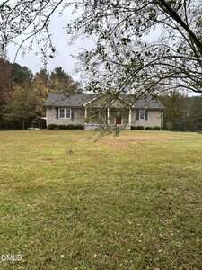 A single-story beige house with white trim and a red front door sits in the center of a large grassy yard at 3105 Tump Wilkins Road in Stem, surrounded by trees under an overcast sky. Branches partially frame the view in the foreground.