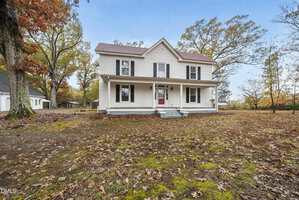 A white two-story house with a covered front porch, red front door, and black shutters sits on a large yard covered with fallen autumn leaves. The house is surrounded by tall trees.