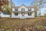 A charming two-story white house in Oxford with a covered front porch, black shutters, and a red door sits among autumn trees and fallen leaves. A detached white garage is visible to the left at 5011 NC 96.