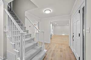 A carpeted staircase with white railings leads upward from a bright entryway featuring light wood floors, white walls, and white doors, with an adjacent open room visible.