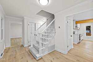 Interior view of a modern home showing a carpeted staircase with white railing, light wood flooring, white walls, and doors leading to other rooms including a visible kitchen with stainless steel appliances.