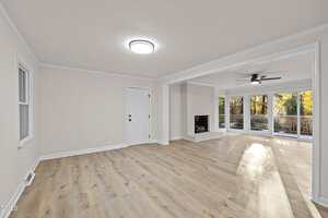 A spacious living room with light wood floors, white walls, a white front door, a fireplace, large windows, and glass doors leading to a deck with trees visible outside. The room has ceiling lights and crown molding.