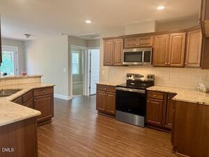 A modern kitchen with wooden cabinets, granite countertops, stainless steel appliances including a stove and microwave, tile backsplash, and wood-look flooring. An adjoining room and hallway are visible in the background.