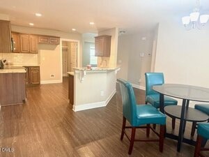 Open kitchen and dining area with wood floors, brown cabinets, granite countertops, a kitchen island, and a round glass dining table with four teal chairs under a chandelier. Neutral wall colors and recessed lighting.