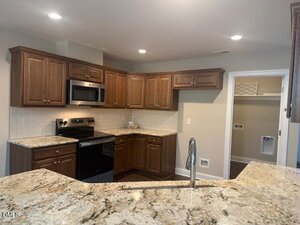 Modern kitchen with brown wooden cabinets, granite countertops, stainless steel microwave, black stove, white tile backsplash, and a gooseneck faucet. A laundry room with hookups is visible through an open doorway.