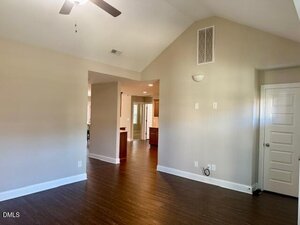 An empty room with beige walls, dark wood flooring, a ceiling fan, and an open doorway leading to a kitchen area. There are visible electrical outlets and a white door on the right wall.