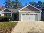 Charming single-story home at 126 Kipling Drive, Oxford, with light gray siding, brown shingle accents, covered front porch, double garage doors, concrete driveway, small front yard, and trees in the background under a clear blue sky.