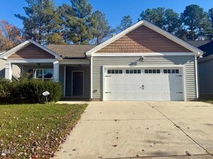 Charming single-story home at 126 Kipling Drive, Oxford, with light gray siding, brown shingle accents, covered front porch, double garage doors, concrete driveway, small front yard, and trees in the background under a clear blue sky.