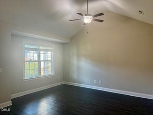 A bright, empty room with dark hardwood floors, light-colored walls, a large window, and a ceiling fan with a light. Sunlight enters through the window, illuminating the space.