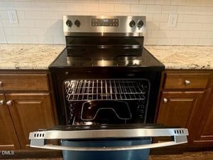 A stainless steel electric stove with four burners and an open oven door. The oven interior is empty except for two racks and a broiler pan. The appliance is set between wooden cabinets on a granite countertop.