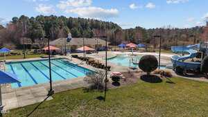 Outdoor aquatic facility with a rectangular lap pool, a separate pool with a blue water slide, several red and blue shade umbrellas, a clubhouse building, and surrounding trees under a partly cloudy sky.