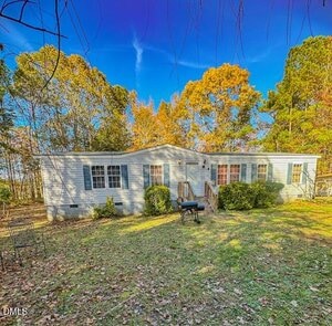 A single-story light gray manufactured home with white trim, front steps, and small bushes sits on a grassy yard with scattered leaves, surrounded by trees under a bright blue sky. A black grill is in front of the house.