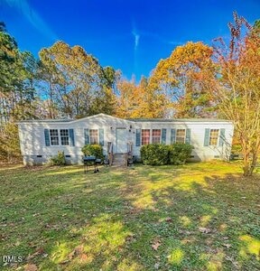 A single-story white manufactured home with blue shutters sits on a grassy lawn at 164 Ramblewood Lane in Henderson, surrounded by trees with green and autumn-colored leaves under a clear blue sky.