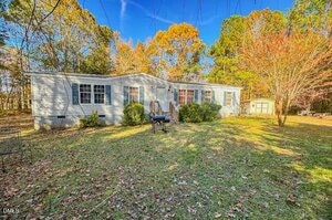 A single-story white manufactured home with multiple windows and a small front porch sits on a grassy yard with scattered leaves, surrounded by trees with autumn foliage. A black grill is positioned near the steps.