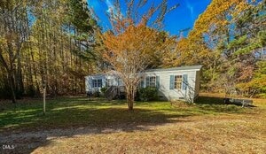 A single-story white manufactured home sits in a clearing surrounded by trees with autumn foliage. A leafless tree stands in the front yard, and a dirt driveway leads up to the house.